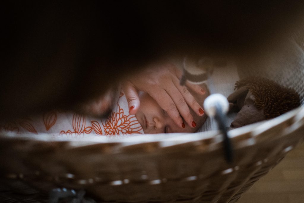 ein Baby liegt in einem Korbbettchen, sein Körper ist mit einer weiß/ rangen Decke eingehüllt, die Mutter des Baby hält ihre Hand auf den Kopf des Baby, das Baby schläft