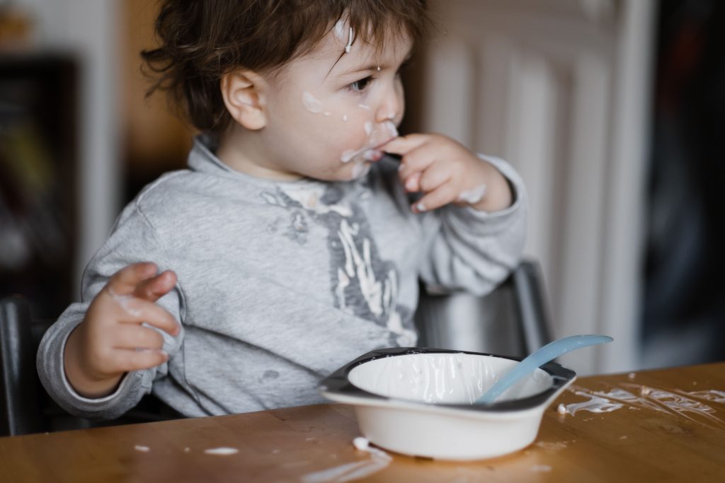 an einem Küchentisch sitzt ein Kleinkind, vor dem Kind steht eine kleine Schüssel, in der Joghurtreste sind , das Kind hält einen Finger in den Mund, an dem Pullover, den das Kind trägt, ist Joghurt, sowie auf dem Tisch vor dem Kind, das Gesicht des Kindes ist ebenfalls voller Joghurt
