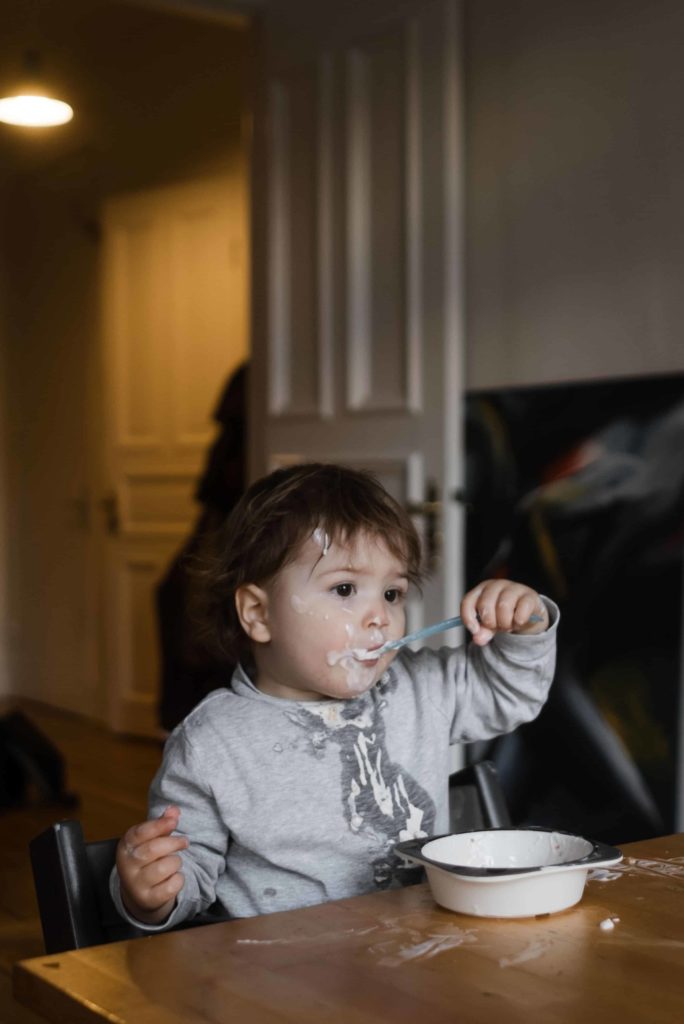 Familienbilder. Ein Kleinkind sitzt an einem Küchentisch, es hält sich einen kleinen Löffel in den Mund, vor dem Kind steht eine Schüssel mit Joghurt, das Gesicht des Kindes ist voll mit Joghurt, sowie der Tisch