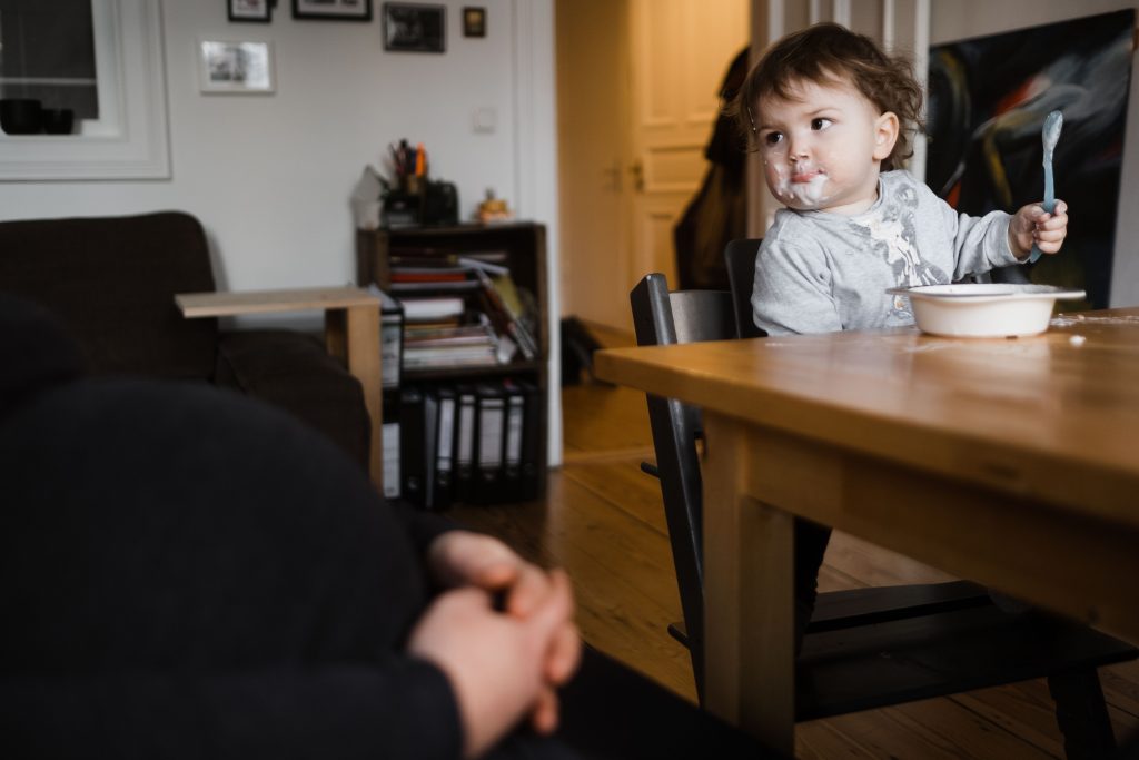 an einem Küchentisch sitzt ein Kleinkind, welches einen Löffel in der Hand hält, vor dem Kind steht eine Schüssel mit Joghurt, das Kind hat einen mit Joghurt verschmierten Mund, es schaut zu derschwangeren Mutter, die neben dem Kind sitzt
