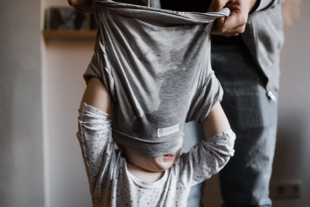 Familienbilder. Einem Kleinkind wird der dreckige Pullover über den Kopf gezogen, das Kind hebt die Arme dabei, sein Gesicht ist mit dem Pullover verdeckt, hinter dem Kind steht der Vater, der dem Kind den Pullover auszieht