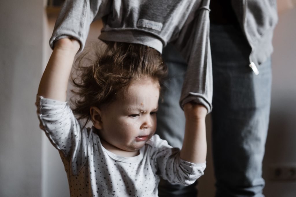 einem Kleinkind wird der dreckige Pullover über den Kopf gezogen, das Kind hebt die Arme dabei, man sieht das Gesicht des Kindes, es schaut nicht begeistert, hinter dem Kind steht der Vater, der dem Kind den Pullover auszieht