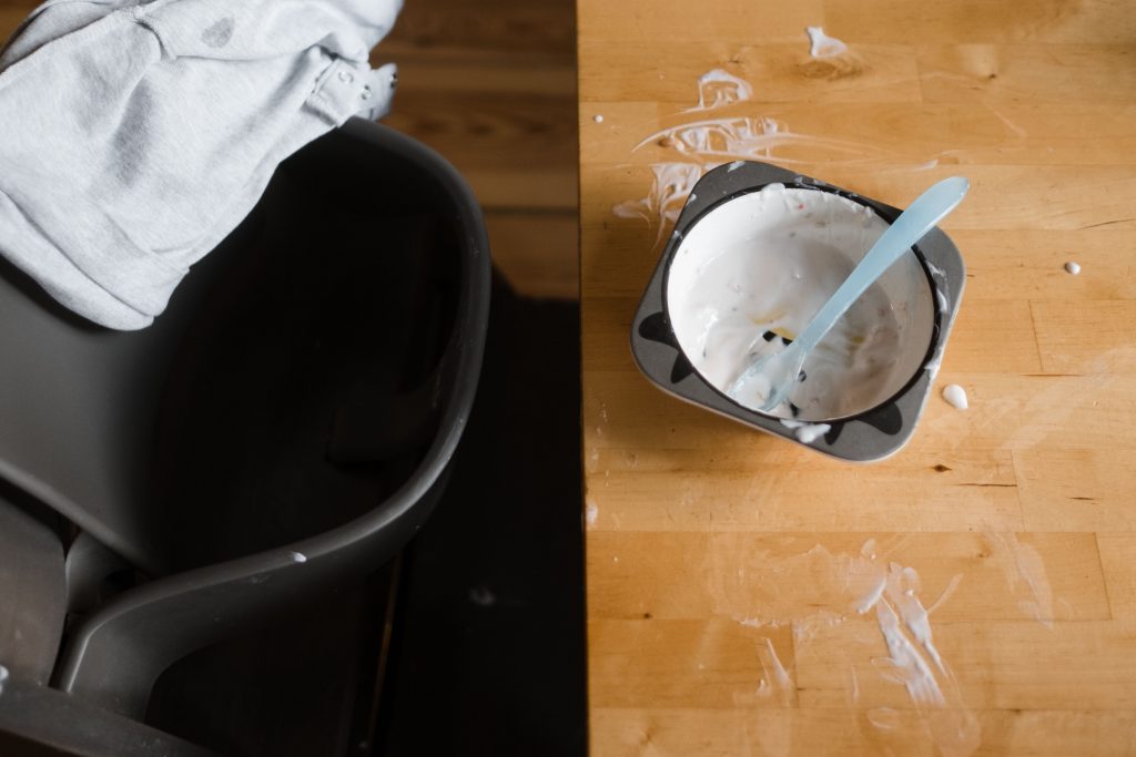 auf einem Küchentisch steht eine leere Schüssel, in der Joghurtreste zu sehen sind, auf dem Tisch sind ebenfalls Joghurtreste, an dem Tisch steht ein Hochstuhl, über der Rückenlehne liegt ein auf linksgedrehter Pullover