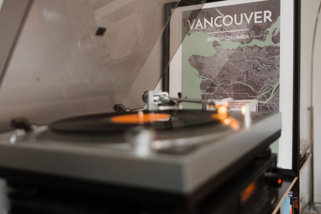 in einem Wohnzimer steht ein offener Plattenspieler, auf dem eine Platteliegt, hinter dem Plattenspieler steht ein Bild, angelehnt an einer Wand, auf dem ein Teil von Vancouver zu sehen ist