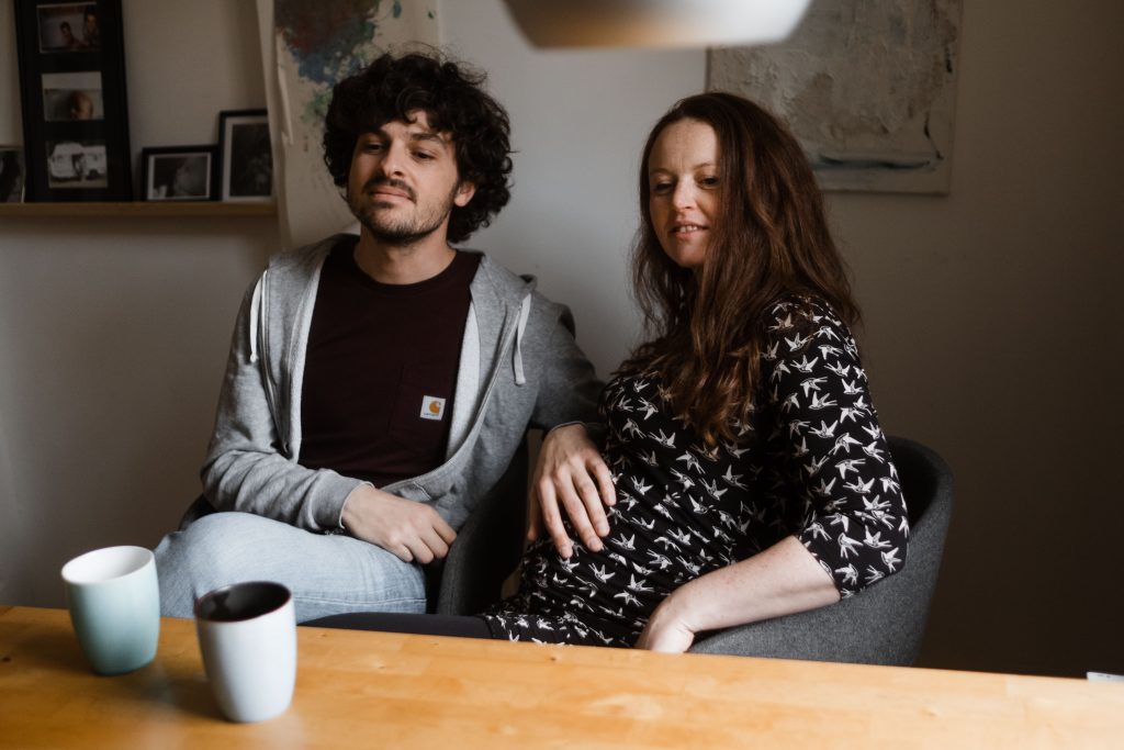 ein Mann und eine schwangere Frau sitzen an einem Küchentisch, vor ihnen stehen 2 Tasse, der Mann legt seine Hand auf den Bauch der Frau, sie schauen in die gleiche Richtung über den Küchentisch