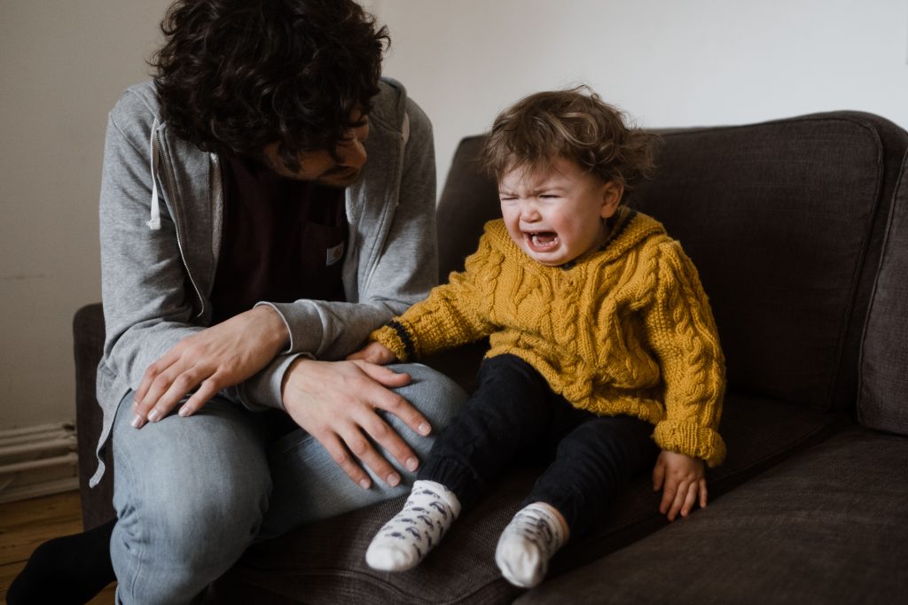 ein Kleinkind sitzt weinend auf einem Sessel im Wohnzimmer, neben ihm sitzt dein Vater, er versucht, das Kind zu beruhigen, das Kind trägt einen gelben, gestrickten Pullover