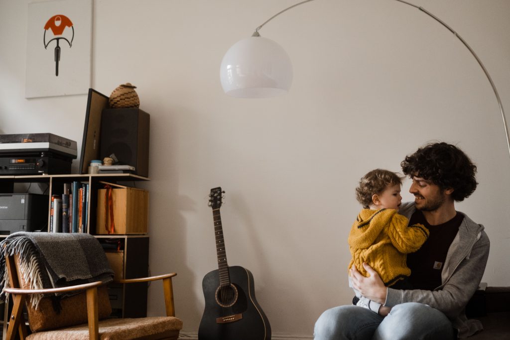 ein Vater sitzt mit seinem Kleinkind auf einem Sofa in einem Wohnzimmer, der Vater hat das Kind auf dem Schoß und schaut es ins Gesicht, über ihnen ist eine gebogen Lampe, neben ihnen steht eine Gitarre, angelehnt an der Wand und ein Bücherregal