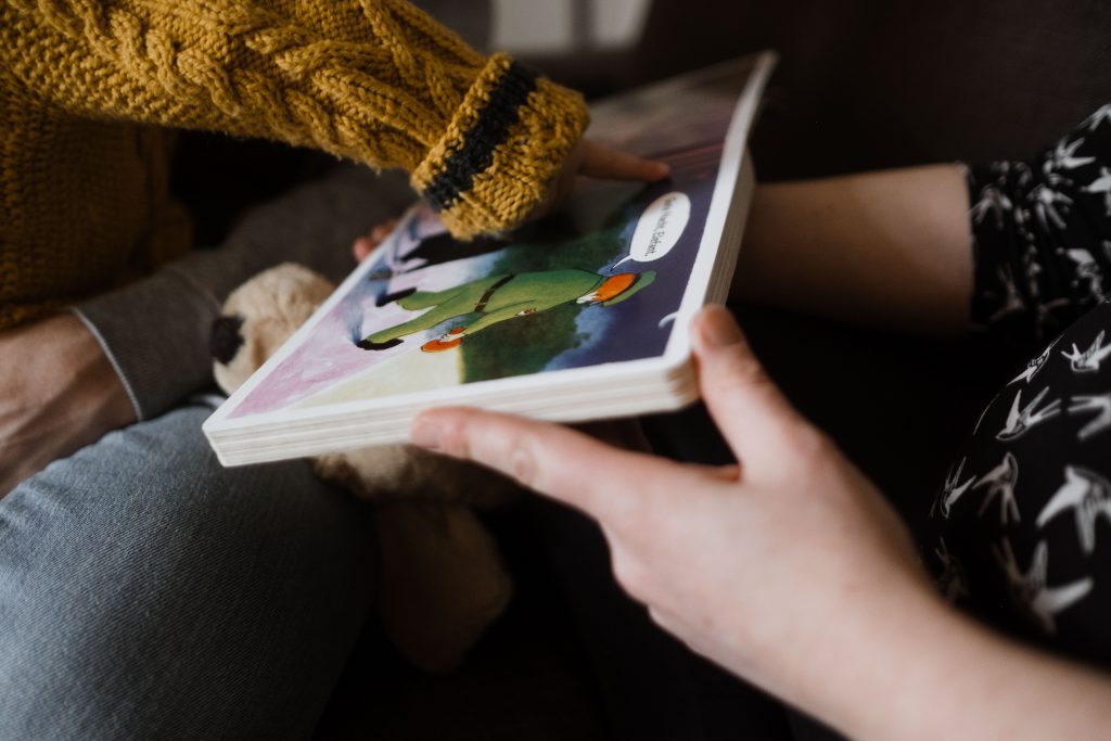 die Mutter hält ein Bilderbuch in der Hand, sie zeigt es in die Richtung ihres Kindes, das Kind zeigt mit dem Finger auf ein Bild in dem aufgeschlagenen Buch