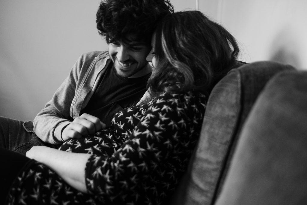 ein Mann und eine Frau sitzen auf einem Sessel im Wohnzimmer, der Mann hält seine Hand auf den Bauch der Frau, sie ist schwanger, ihre Köpfe halten sie aneinander, der Mann schaut auf den Bauch der Frau