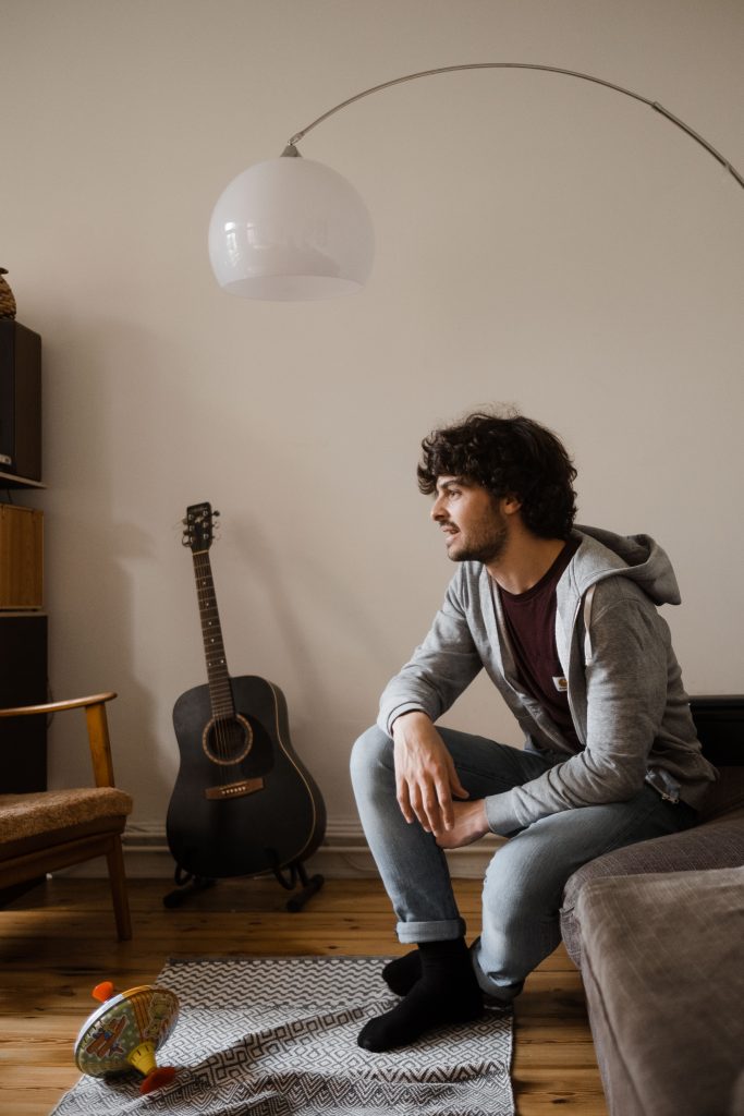 ein Mann sitzt auf einem Sessel im Wohnzimmer, er stützt sich an seinem Knie ab und schaut in die Ferne, vor ihm liegt ein kleiner Teppich, auf dem ein Brummkreisel liegt, über ihm steht eine gebogene Lampe, neben ihm steht eine Gitarre, die an der Wand gelehnt ist