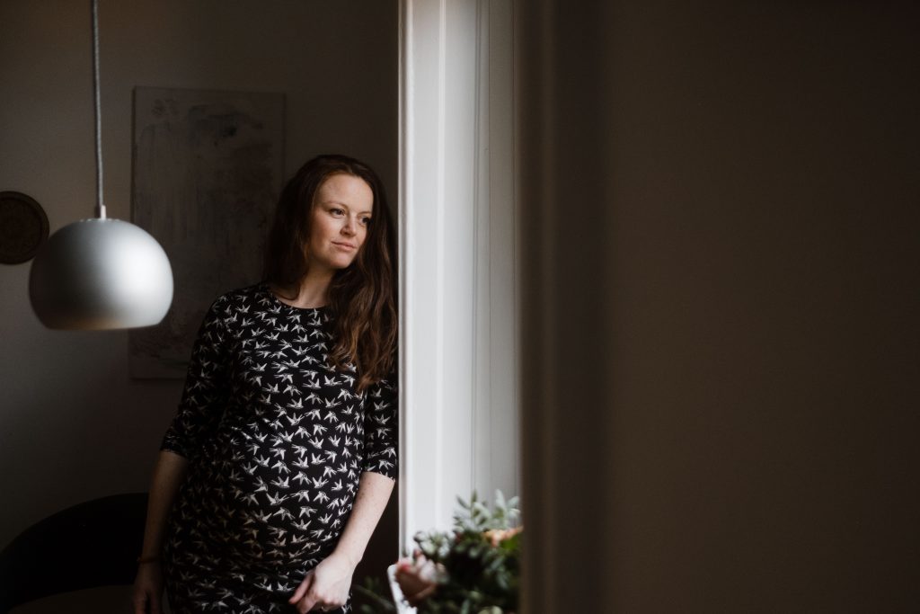 eine schwangere Frau steht angelehnt an einem Fensterrahmen, sie schaut verträumt hinaus, auf der Fensterbank steht ein Blumenstrauß, neben ihr hängt eine runde Deckenlampe