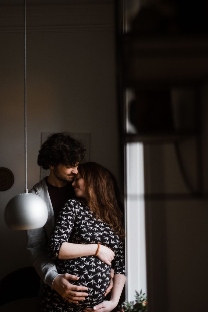 ein Mann und eine Frau stehen angelehnt an einem Fensterrahmen, der Mann hält seine Hände um die schwangere Frau und hält die Hände an ihren Bauch, die Frau legt ihren Kopg an den Hals des Mannes, der Mann schaut auf das Gesciht der Frau