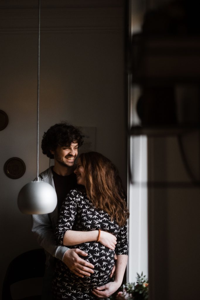 ein Mann und eine Frau stehen angelehnt an einem Fensterrahmen, der Mann hält seine Hände um die schwangere Frau und hält die Hände an ihren Bauch, die Frau legt ihren Kopg an den Hals des Mannes, der Mann lacht