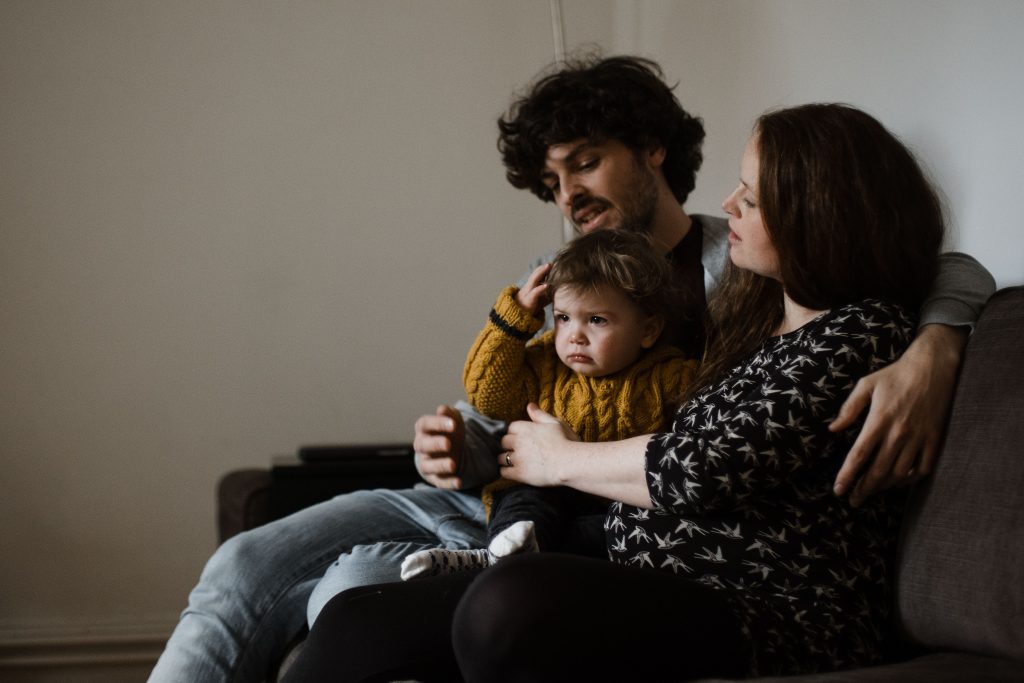 ein Mann und eine Frau sitzen mit ihrem Kleinkind auf einem Sofa im Wohnzimer, das Kind hebt die Arme hoch, es schaut quengelig, die Fra legt hren Arm um den Bauch des Kindes
