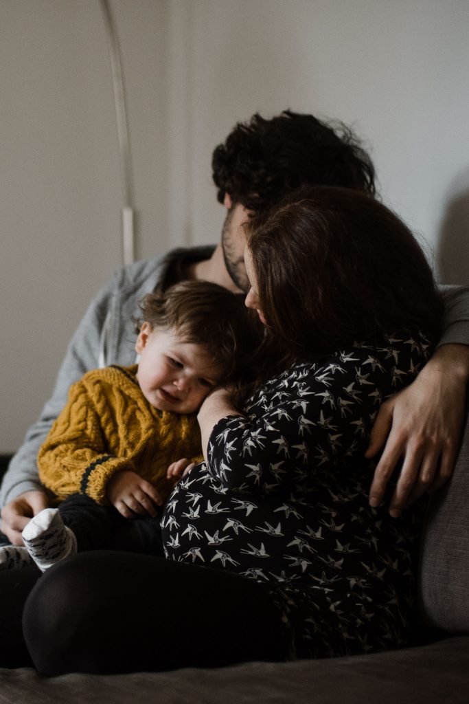 ein Mann und eine Frau sitzen mit ihrem Kleinkind auf einem Sofa im Wohnzimmer, die Frau ist schwanger, sie hält ihre Hand an das Gesicht des Kindes, der Mann hat das Kind auf dem Schoß, er hält seine Hand um die Schulter der Frau, das Kind ist quengelig
