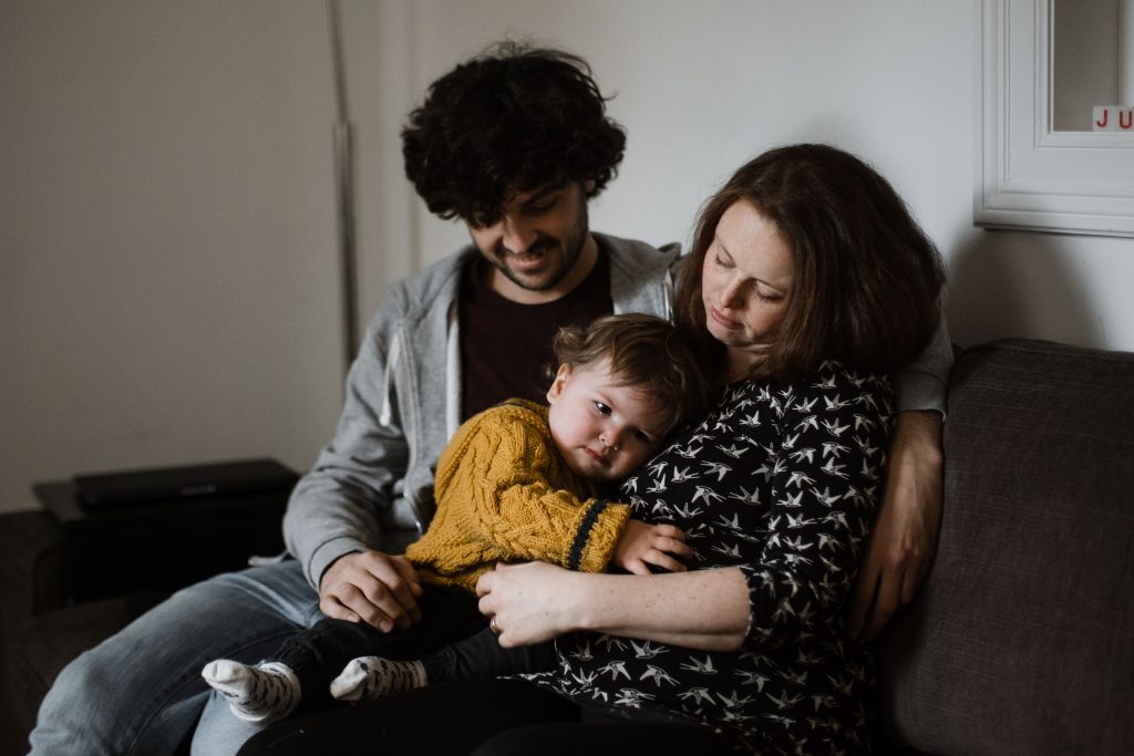 ein Mann und eine Frau sitzen mit ihrem Kleinkind auf einem Sofa im Wohnzimer, das Kind lehnt sich an den Bauch der Mutter, sie ist schwanger, der Mann sitzt neben der Frau, er hält seine Hand um das Kind, das kind hat Tränen in den Augen, der Mann und die Frau schauen beide auf das Kind