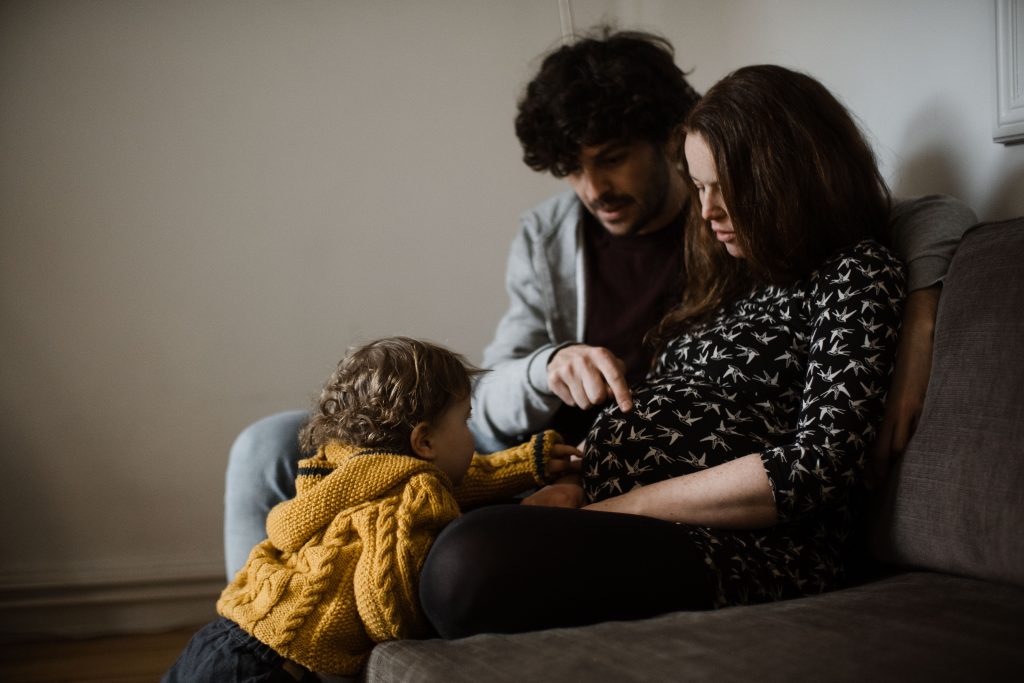 ein Mann und eine Frau sitzen auf einem Sofa nebeneinander, vor ihnen steht ihn Kleinkind, es lehnt sich an die Beine der Frau, der Mann hält einen Finger auf den Bauch der Frau, sie ist schwanger, der Mann und die Frau schauen beide auf das Kind