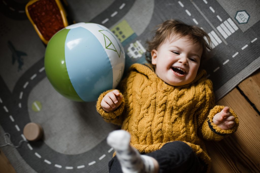 ein Kleinkind liegt mit dem Rücken auf einem Spielteppich, auf dem Straßen sind, es lacht und hebt dabi die Beine hoch, das Kind trägt einen gelben Strickpullover, neben dem Kind liegt ein aufgeblasener Gummiball