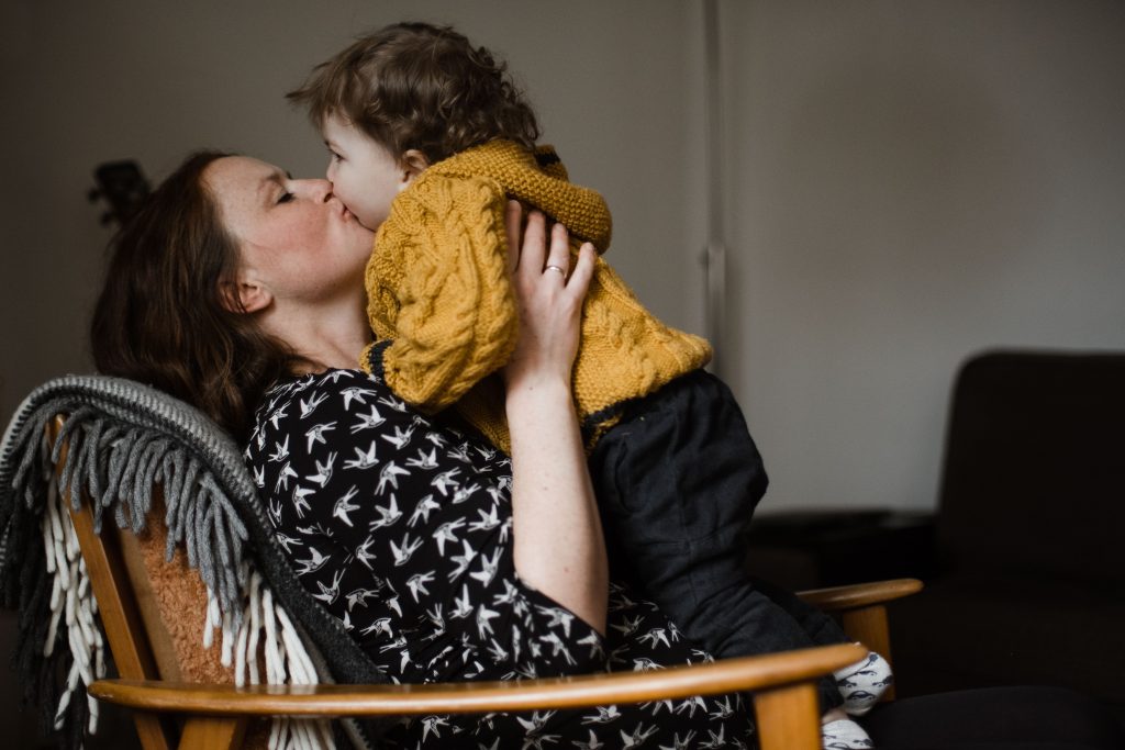 eine schwangere Frau sitzt auf einem Holzsessel, in ihren Armen hält sie ein Kleinkind, sie küsst es auf den Mund und schließt dabei die Augen