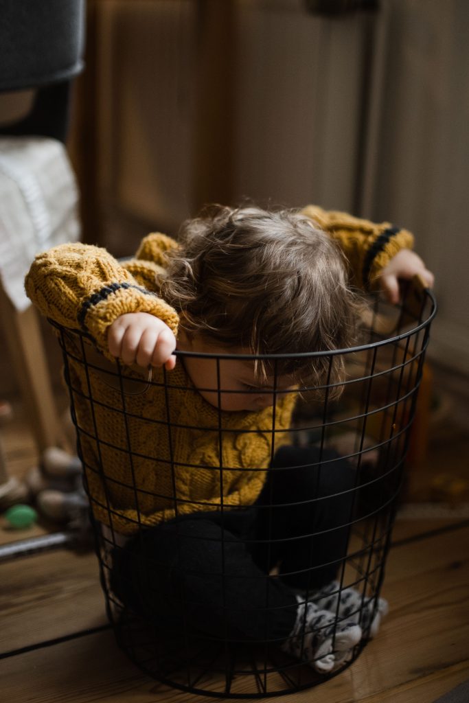 in einem Drahtkorb sitzt ein Kleinkind, es trägt einen gelben Strickpullover, es stützt sich mit den Händen am Rand des Korbes und schaut dabei auf den Boden des Korbes