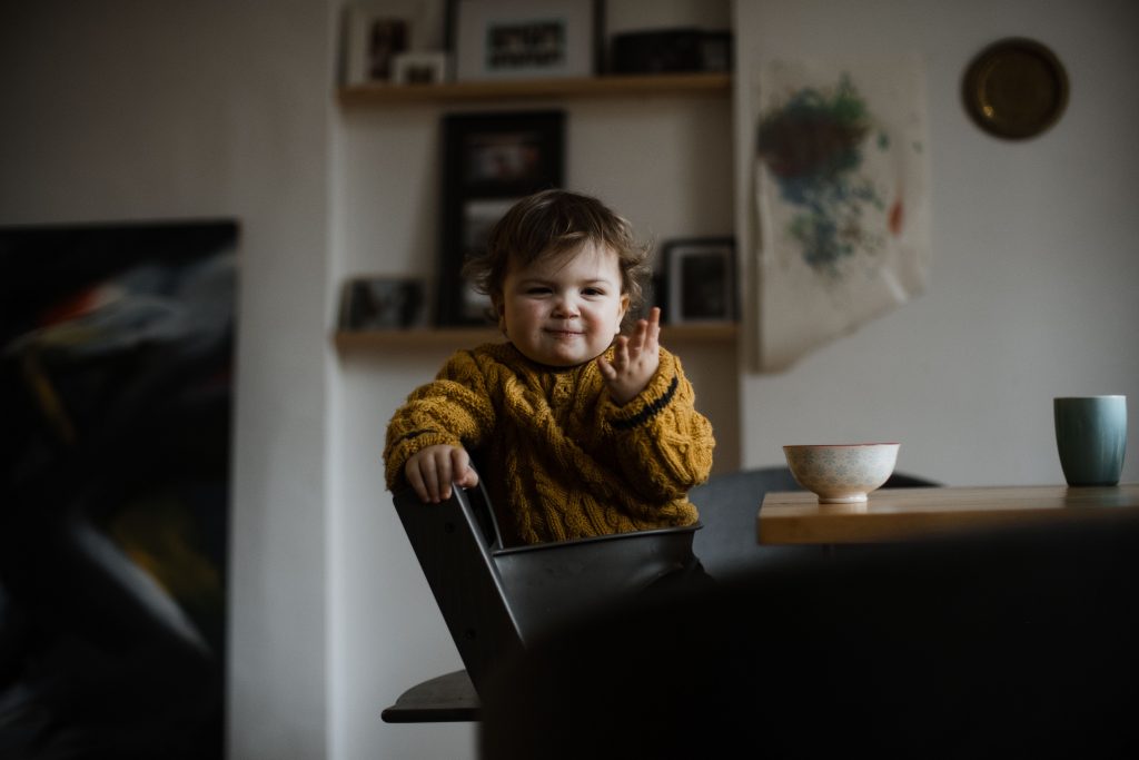 ein Kleinkind sitzt auf einem Hochstuhl an einem Küchentisch, vor ihm steht eine kleine Schüssel, das Kind hebt die Hand hoch