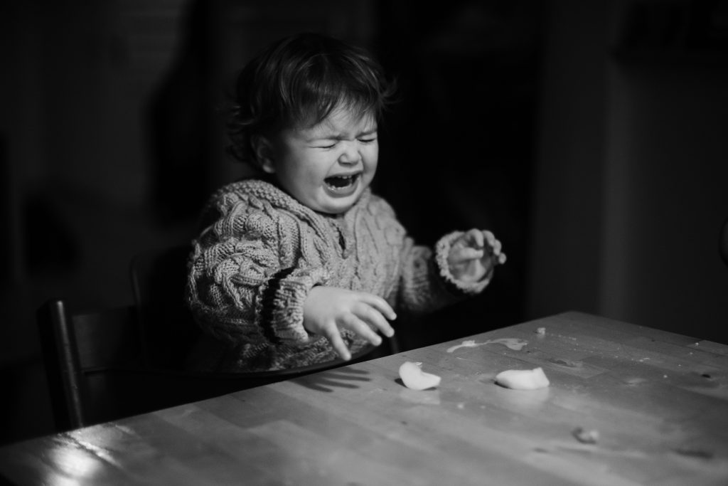 ein Kleinkind sitzt an einem Küchentisch, vor dem Kind auf dem Tisch liegen Apfelstücke, das Kind hält die Hande hoch und weint