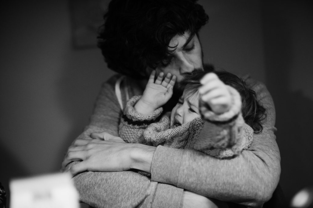 ein Vater hält sein Kleinkind in dem Armen, das Kind hält seine Arme hoch, es verzieht das Gesicht, der Mann küsst dem Kind uf den Kopf