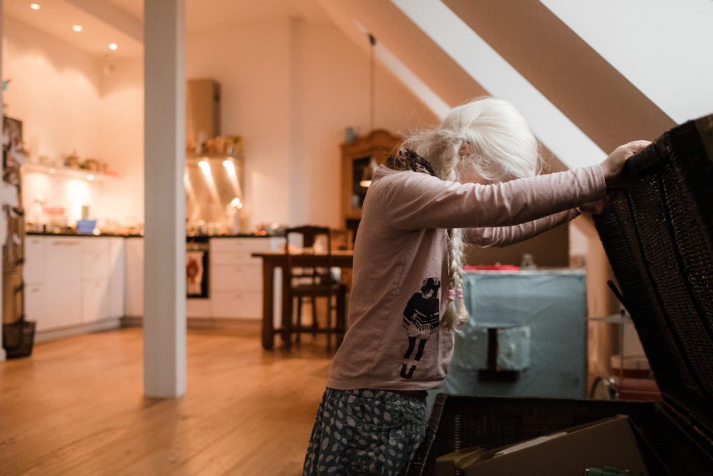 ein Mädchen steht in einem Wohnzimmer, sie beugt sich über eine goße Kiste, die sie geöffnet hält, hinter ihr ist die offene Küche zu sehen , sowie Dachschrägen, in denen Fenster eingesetzt sind, das Mädchen trägt einen Rock und einen Pullover, sowie 2 Zöpfe
