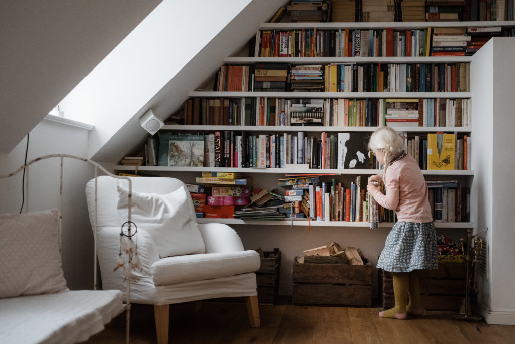 vor einem hohen Bücherregal in einem Wohnzimmer steht ein Mädchen, sie trägt einen Rock und einen Pulver, sie ist blond und hat 2 Zöpfe, sie zieht 4 Bücher aus dem Bücherregal, das Regal ist angepasst an einer Dachschräge in dem Wohnzimmer, neben bem Regal steht ein weißes Sofa
