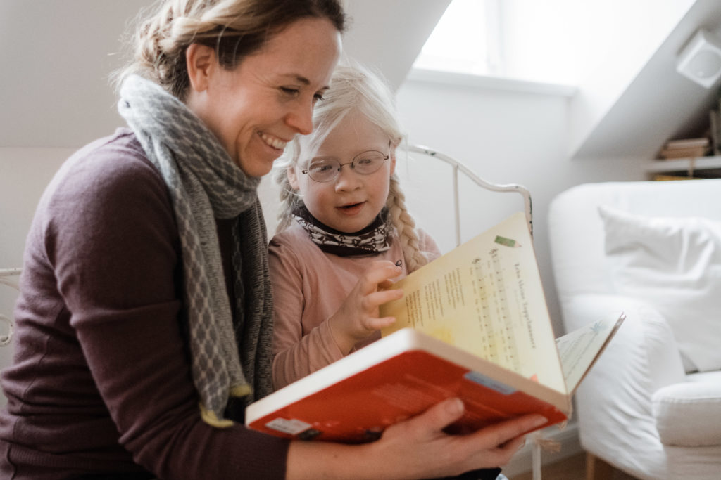 ein Mädchen sitzt mit ihrer Mutter auf einem Sofa in einem Wohnzimmer, die Mutter hat ein Buch aufgeschlagen, beide schauen in das Buch, beide haben ein Lächeln im Gesicht, hinter ihnen ist ene Dachschräge mit einem Fenster, durch das die Sonne hineinstrahlt