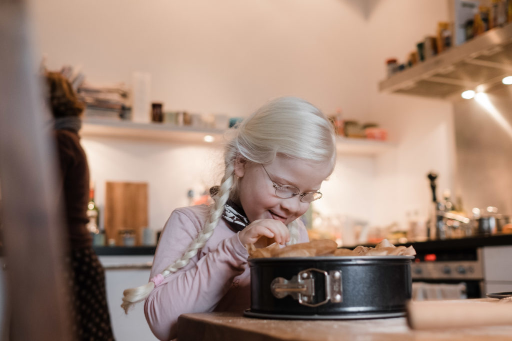 ein Mädchen steht an einem großen Küchentisch, sie schaut in eine Springform, die mit Backpapier ausgelegt ist, sie hat 2 Zöpfe und eine Brille, sie grinst, ihre Hand hält sie in die Springform