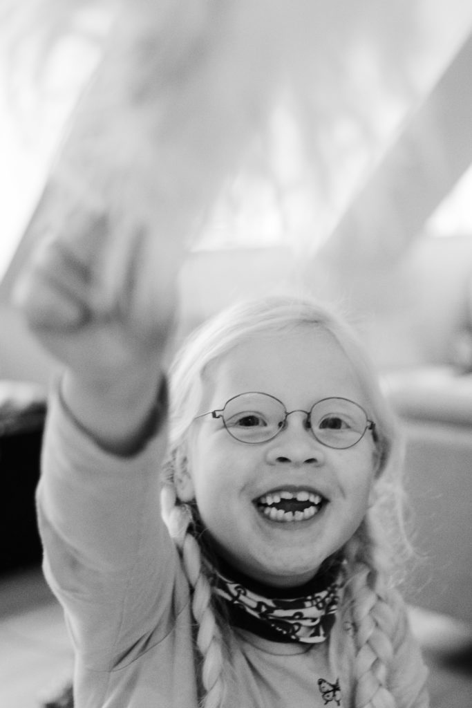 ein Mädchen hält eine große Feder in der Hand, sie hat eine Brille auf und 2 Zöpfe, sie grinst