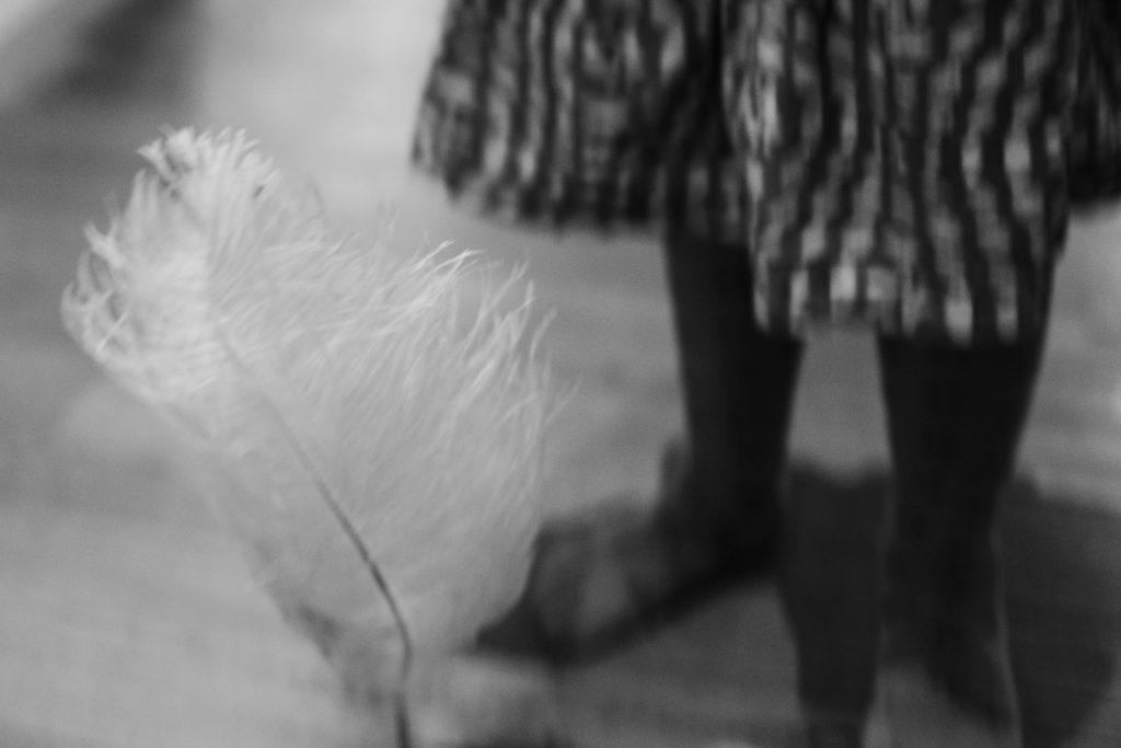 eine große Feder ist im Vordergruns, dahinter steht ein Mädchen, sie trägt einen Rock und eine Strumpfhose