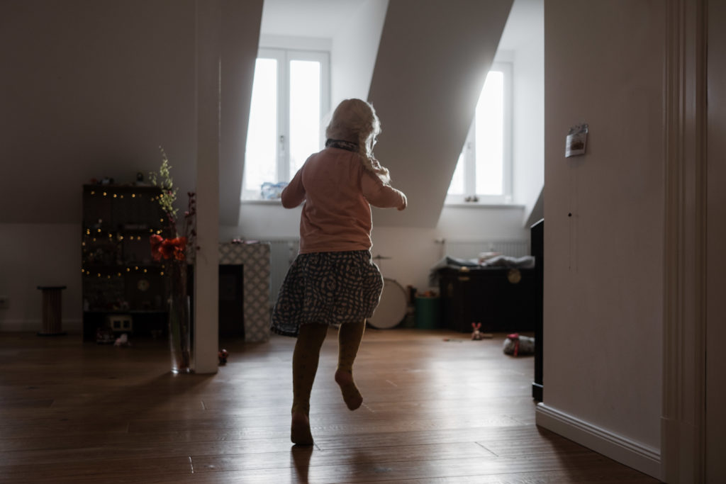 ein Mädchen läuft durch ein großes Wohnzimmer, durch die 2 Fenster scheint die Sonne hinein, das Mädchen trägt einen Rock und einen Pullover