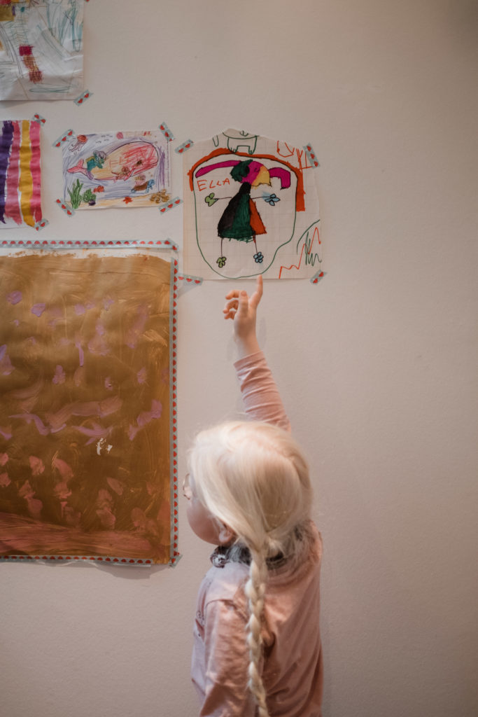 ein Mädchen zeigt mit ihrem Finger auf ein von ihr gemaltes Bild, dass neben anderen gmalten Bilder an einer Wand hängt