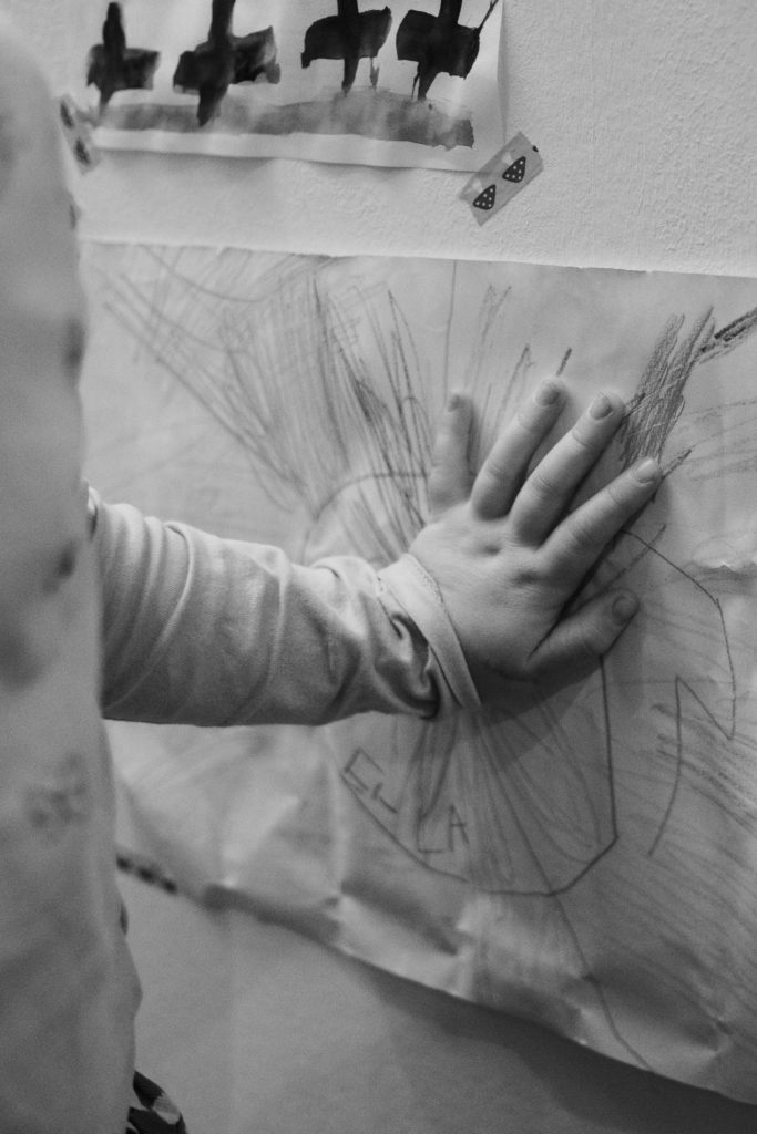 ein Mädchen hält ihre Hand auf ein von ihr gemaltes Bild, dass an einer Wand hängt