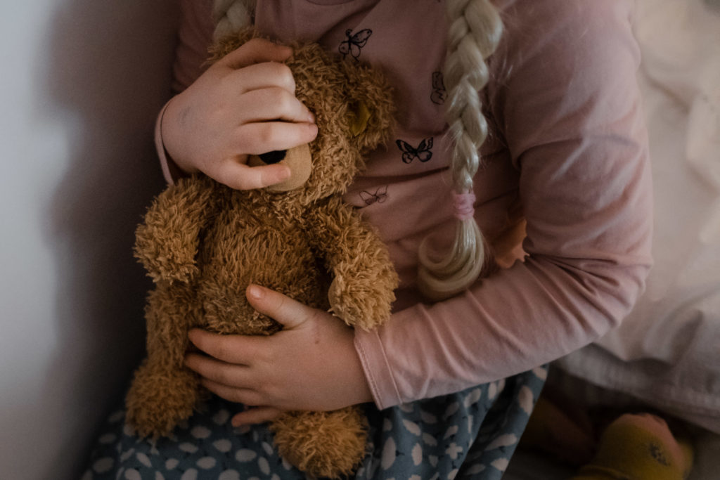 ein Mädchen hält einen braunen Teddybären in ihren Händen, sie trägt einen Rock, einen Pullove und 2 Zöpfe