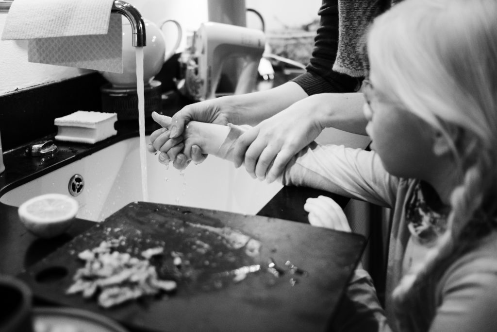eine Mutter steht mit ihrer Tochter an einem Waschbecken in der Küche, der Hahn läuft, die Mutter hält den Arm ihrer Tochter unter den Waserstrahl, am Waschbeckenrand steht ein benutztes Schneidebrett