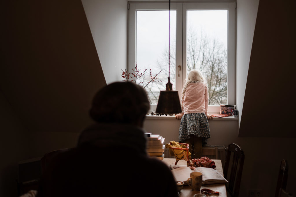 ein Mädchen steht an einem Fenster, sie schaut hinaus, hinter ihr steht ein großer Küchentisch, an dem ihre Mutter sitzt mit Blick auf ihre Tochter