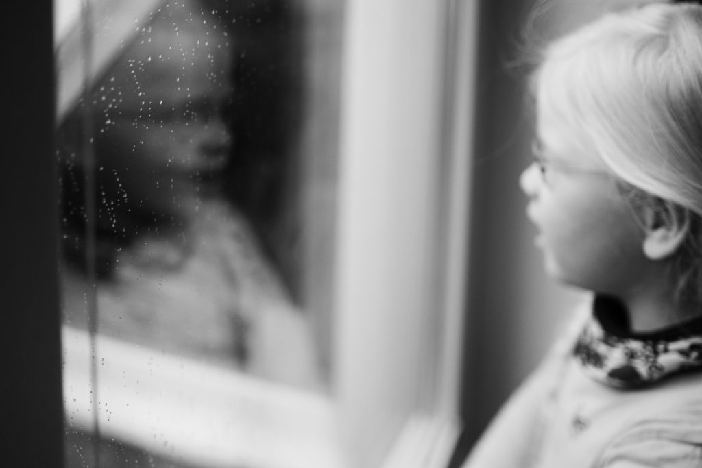 ein Mädchen steht vor einem Fenster, an dem Fenster sind Regentropfen,sie spiegelt sich in dem Fenster