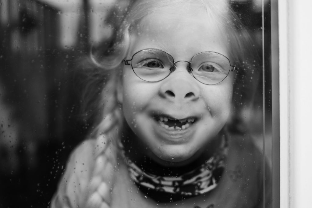 ein Mädchen mit Brille und 2 Zöpfen drückt ihr Gesicht gegen eine Fensterscheibe, sie hat dadurch eine platte Nase