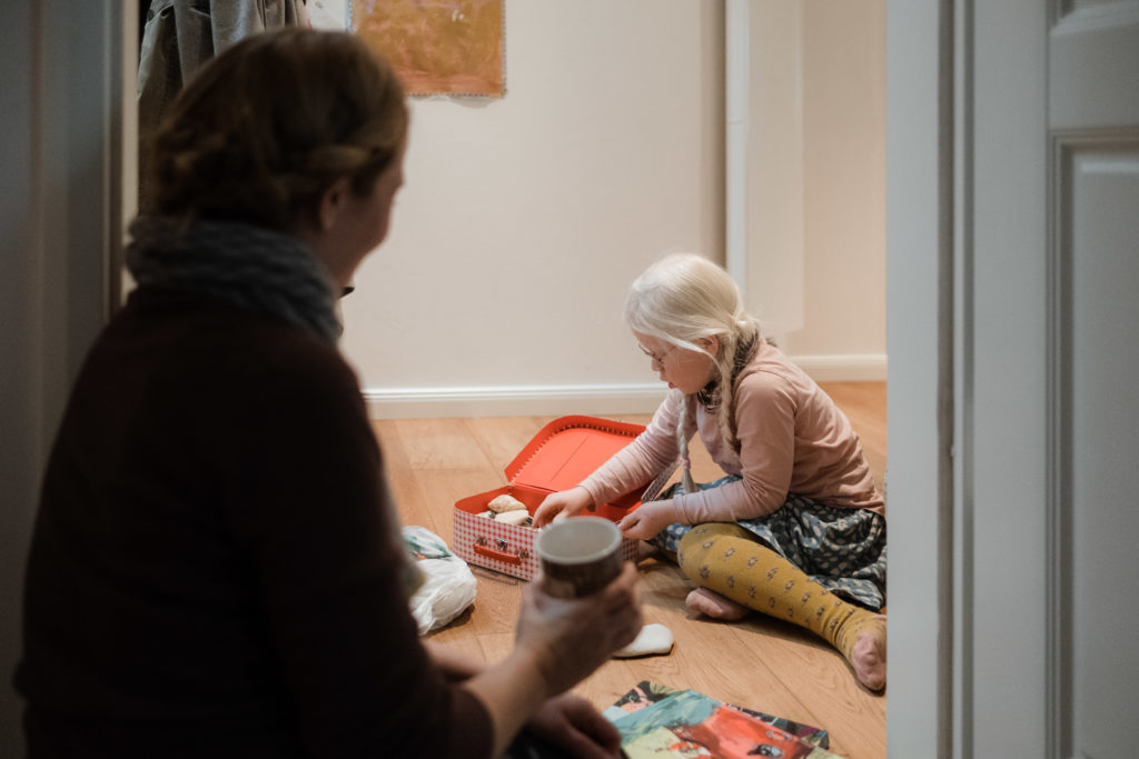 ein Mädchen sitzt auf dem Holzfußboden in einem Wohnzimmer, sie packt einen kleinen, roten Spielzeugkoffer aus, bei ihr sitzt ihre Mutter, sie hält eine Tasse in der Hand und schaut auf ihre Tochter