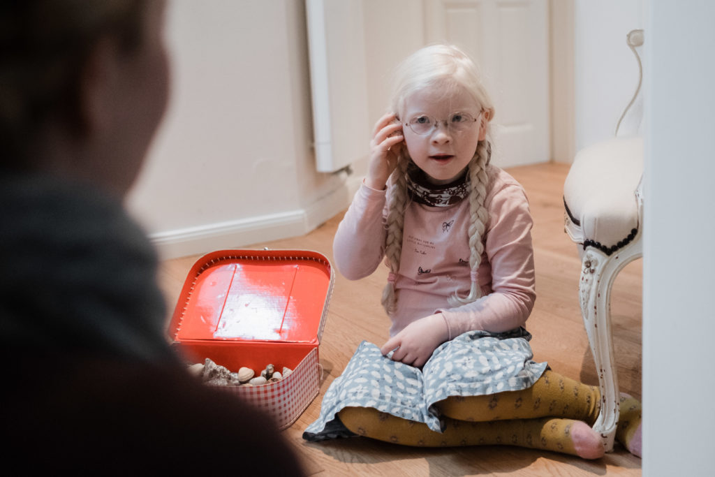 ein Mädchen sitzt auf dem Wohnzimmerfußboden, neben ihr liegt ein kleiner, roter Spielzeugkoffer, das Mädchen trägt einen Rock, einen Pullover, eine Brille und 2 Zöpfe, mit der rechten Hand fasst sie sich an das Ohr, sie schaut zu ihrer Mutter, die vor ihr sitzt
