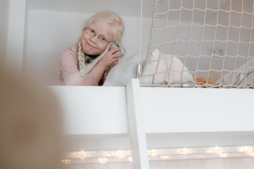 ein Mädchen sitzt auf einem Hochbett, sie trägt 2 Zöpfe und eine Brille, sie hält sich ein Kuscheltier an die Wange und grinst