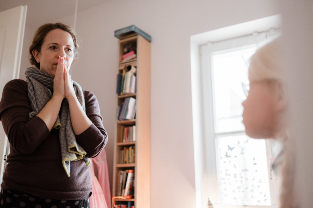 eine Mutter steht vor ihrer Tochter in einem Wohnzimmer, sie legt ihre Hände vor sich und hält sie vor ihren Mund, sie schaut zu ihrer Tochter und überlegt, die Tochter schaut zu der Mutter