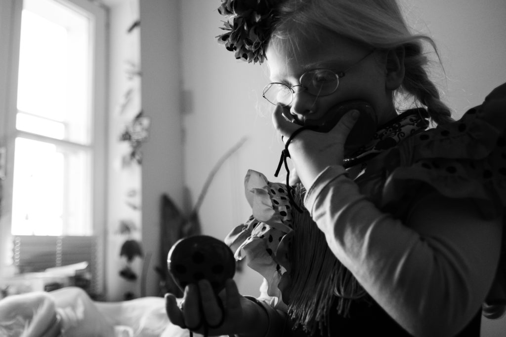 ein Mädchen trägt ein Kleid und ein Haarreif und eine Brile, sie hält eine kleinen Ball in der Hand, mit der anderen Hand hält sie sich vor den Mund