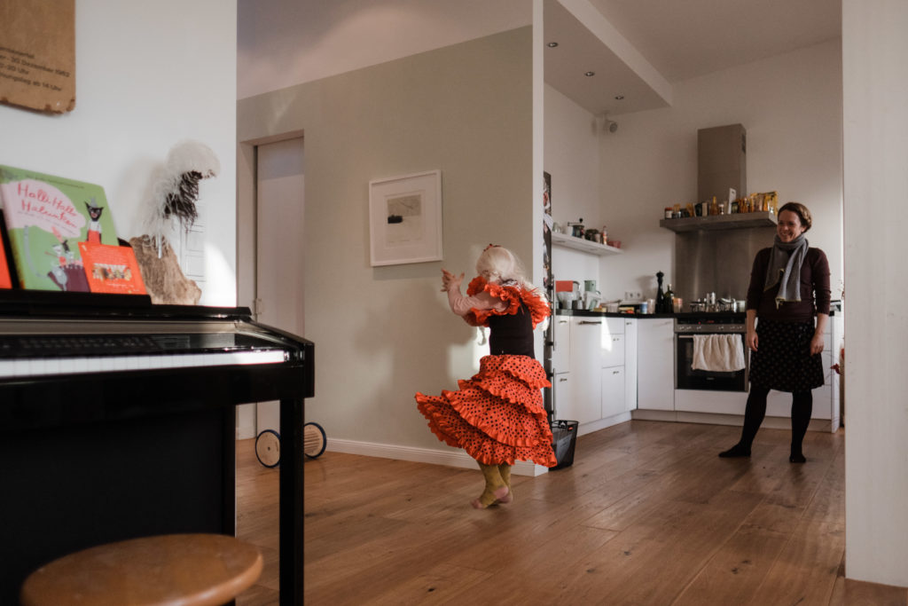 in einem Wohnzimmer tanzt ein Mädchen,sie trägt ein rotes Kleid und einen Haarreif ( sie ist verkleidet), ihre Mutter steht in der ofenen Küche und beobachtet sie