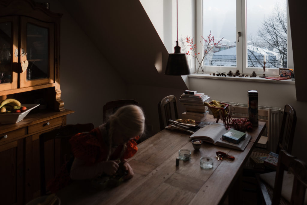 an einem großen Küchentisch sitzt ein Mädchen, sie beugt sich auf den Tisch und schaut auf den Tisch, am Ende des Tich ist ein Fenster, durch das Licht hineinfällt