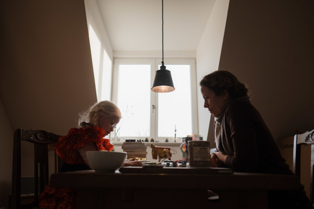 eine Mutter sitzt mit ihrer Tochter an einem großen Küchentisch, sie sitzen sich gegenüber, am Kopf des Tiches ist ein Fenster, durch das Licht in die Küche fällt