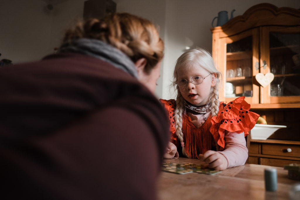 eine Mutter sitzt mit ihrer Tochter an einen großen Küchentisch, sie sitzen sich gegenüber, ein Spiel steht zwischen ihnen auf dem Tich, die Tochter schaut zur Mutter, hnter der Tochter steht ein Holzschrank der von der Sonne angestraht wird